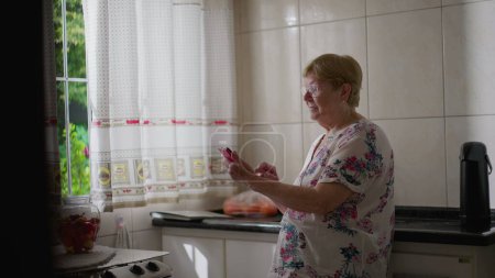 Téléchargez les photos : Candid femme âgée en utilisant un appareil smartphone à la maison appuyé sur l'évier. Style de vie domestique de dame âgée engagée avec la technologie moderne - en image libre de droit