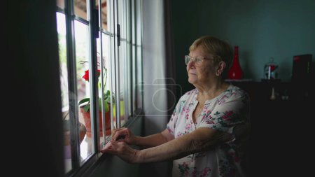 Téléchargez les photos : Femme âgée avec l'expression pensive tenant la barre de fenêtre, scène contemplative de la vieillesse authentique style de vie domestique - en image libre de droit