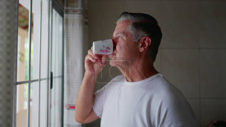 Photo for Contemplative middle-aged man standing by kitchen window sipping coffee during morning ritual, domestic everyday lifestyle of gray hair man sips warm drink - Royalty Free Image