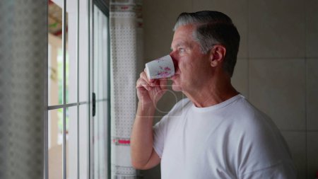 Photo for Contemplative middle-aged man standing by kitchen window sipping coffee during morning ritual, domestic everyday lifestyle of gray hair man sips warm drink - Royalty Free Image