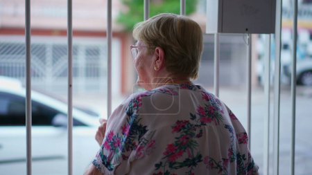 Téléchargez les photos : Femme âgée debout à l'intérieur de la maison fermée, surplombant le trottoir, s'accrochant à une barre métallique, observant le quartier - en image libre de droit