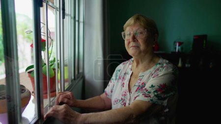 Photo for Portrait of an elderly woman looking at camera standing by window at home. Authentic domestic old age lifestyle of senior person in 80s - Royalty Free Image
