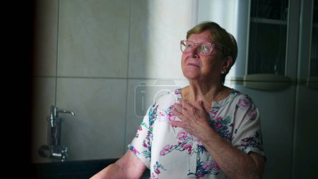 Foto de Feliz anciana pensativa de pie junto a la ventana en la cocina. Pensativo mayor mujer persona en los años 80 soñando despierto y contemplando la vida - Imagen libre de derechos