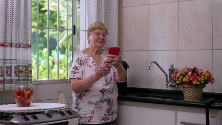Téléchargez les photos : Femme âgée parlant avec un membre de la famille sur vidéo tout en tenant le téléphone et debout dans la cuisine de la maison par la fenêtre. Dame âgée utilisant la technologie moderne pour communiquer avec les parents - en image libre de droit
