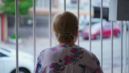 Foto de Mujer mayor de pie dentro de una casa cerrada, con vistas a la acera, aferrándose a la barra de metal, observando el vecindario - Imagen libre de derechos