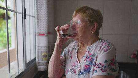 Téléchargez les photos : Femme âgée pensive buvant du café près de la cuisine de window _ s. Une personne âgée caucasienne regardant l'horizon avec une expression réfléchie réfléchissant solution - en image libre de droit