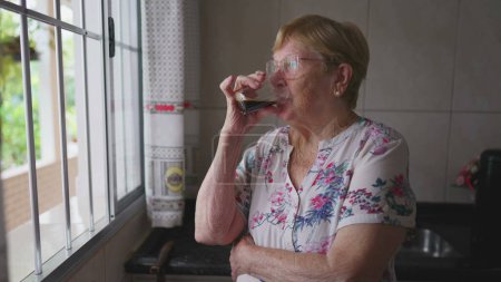 Foto de Una anciana pensativa bebiendo café en la cocina de window _ s. Una persona mayor caucásica de edad avanzada mirando al horizonte con expresión reflexiva ponderando solución - Imagen libre de derechos