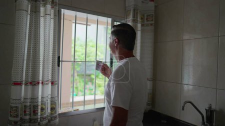 Foto de Contemplación matutina del hombre de mediana edad bebiendo café por la ventana de la cocina, ritual diario doméstico - Imagen libre de derechos