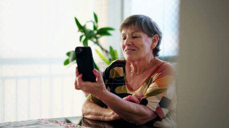 Téléchargez les photos : Candid Senior Woman saluant et tenant le téléphone tout en s'engageant dans la communication vidéo sentiment de connexion avec un membre de la famille en raison de la technologie - en image libre de droit