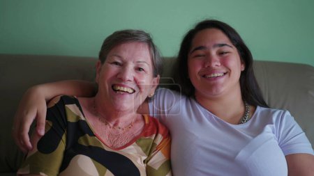 Téléchargez les photos : Portrait de joyeuse et diverse grand-mère et petite-fille sur canapé riant et souriant, Asiatique jeune adulte avec bras autour aîné caucasien - en image libre de droit