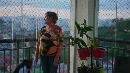 Téléchargez les photos : Femme plus âgée contemplative debout par la fenêtre au balcon de l'appartement regardant à l'extérieur à travers le verre à la vue sur la ville. Solitude thème des personnes âgées dans les années 70 - en image libre de droit