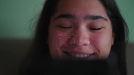 Photo for Joyful Brazilian teen girl react_s positively to content online while holding phone. Close-up of a happy diverse Asian woman_s face smiling reading message - Royalty Free Image