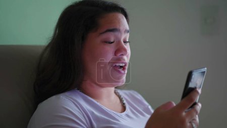 Foto de Happy Brazilian Teen Girl reacciona positivamente al contenido en línea, sosteniendo el teléfono. Escena cándida de una mujer asiática diversa sonriendo mientras lee el mensaje - Imagen libre de derechos