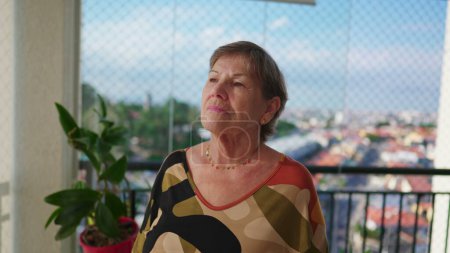 Foto de Mujer mayor reflexiva Reflexionando sobre la vida de pie en el balcón del apartamento mirando a la vista de la ciudad con mirada contemplativa mientras está en contemplación - Imagen libre de derechos