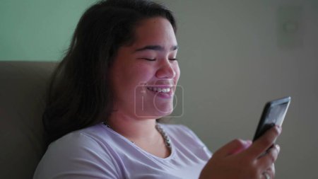 Foto de Happy Brazilian Teen Girl reacciona positivamente al contenido en línea, sosteniendo el teléfono. Escena cándida de una mujer asiática diversa sonriendo mientras lee el mensaje - Imagen libre de derechos