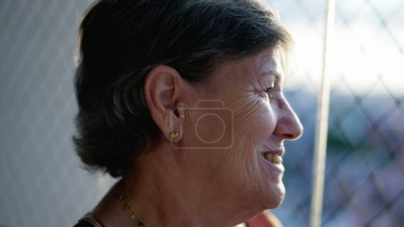 Téléchargez les photos : Contemplative heureuse femme âgée debout par la fenêtre regardant à l'extérieur à travers le verre de fenêtre. Une personne âgée souriante dans les années 70 - en image libre de droit