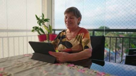 Foto de Mujer mayor feliz usando tableta en casa. Persona femenina madura mayor mirando la pantalla de tecnología moderna en el balcón del apartamento - Imagen libre de derechos