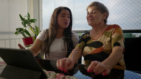 Téléchargez les photos : Joyeuse grand-mère devant l'appareil tablette avec sa petite-fille asiatique diversifiée au balcon de l'appartement. Scène intergénérationnelle d'authentique moment familial diversifié - en image libre de droit