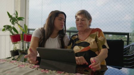 Photo for Intergenerational Bonding Diverse Granddaughter Assisting Caucasian Grandmother with Tablet on Apartment Balcony. Sharing Technological Knowledge in a Generational Family Scene - Royalty Free Image