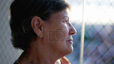 Téléchargez les photos : Profil réfléchi de la femme âgée ridée debout par la fenêtre regardant à travers la fenêtre de verre à la maison. Personnes âgées méditatives et chères dans les années 70 - en image libre de droit