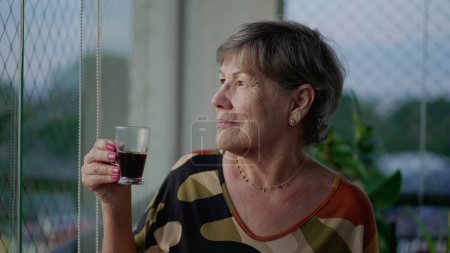 Téléchargez les photos : Une chère femme plus âgée buvant du café tout en contemplant la vue depuis le balcon de l'appartement. Dame aînée réfléchie - en image libre de droit