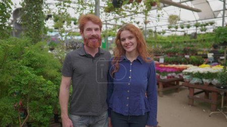 Foto de Una feliz pareja pelirroja sonriendo a la cámara de pie dentro de la floristería. Caucásico pelo rojo Hombre y mujer dentro de tienda local - Imagen libre de derechos