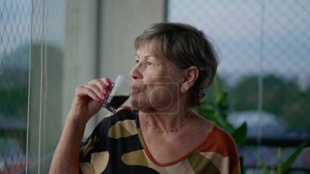 Téléchargez les photos : Une chère femme plus âgée buvant du café tout en contemplant la vue depuis le balcon de l'appartement. Dame aînée réfléchie - en image libre de droit
