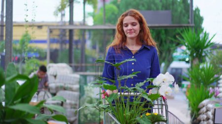 Foto de Mujer alegre de compras de plantas y floresen la tienda de horticultura. Una mujer pelirroja con blusa azul empujando carro caminando por la isla en busca de productos para la compra - Imagen libre de derechos