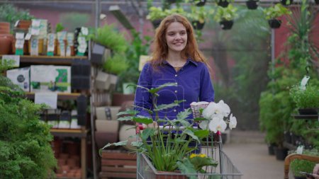 Téléchargez les photos : Happy Woman en marchant dans l'allée du magasin d'horticulture. Femme Shopper avec panier à la recherche d'achats de plantes - en image libre de droit