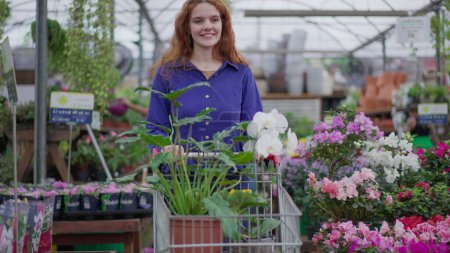 Foto de Mujer alegre seleccionando plantas y flores con carro en una floristería. Compras de clientes femeninos en la tienda local - Imagen libre de derechos