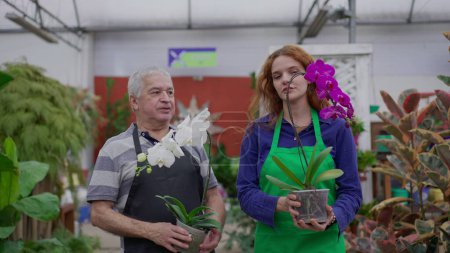 Téléchargez les photos : Employés de l'horticulture marchant avec des fleurs dans l'allée du magasin. Une jeune femme et un homme âgé portant des tabliers et portant des plantes - en image libre de droit