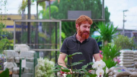 Téléchargez les photos : Jeune homme heureux avec panier explorant l'allée dans un magasin d'horticulture. Homme client achats pour les plantes à la boutique de jardin locale - en image libre de droit