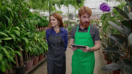 Foto de Joyful Redhaired Staff Colegas explorando la tienda de flores con Tablet. Hombre y mujer verde-proned que abraza la tecnología en negocio local de la planta - Imagen libre de derechos