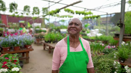 Foto de Una empleada negra mayor caminando en Flower Shop con delantal verde. Una mujer afroamericana paseando por la tienda de plantas - Imagen libre de derechos