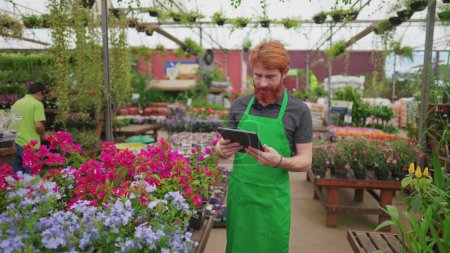 Photo for Young Man in Green Apron Utilizing Tablet for Inventory at Flower Shop. Male Employee Engaging with Modern Tech in Retail Environment - Royalty Free Image