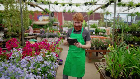 Photo for Smiling Red-Haired Employee with Tablet in Flower Shop. Young Man in Apron Leveraging Modern Tech in Local Business - Royalty Free Image