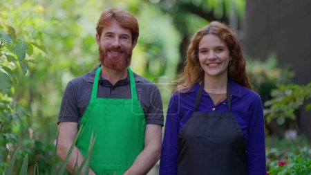 Foto de Hombre y mujer Empleados de horticultura sonriendo a la cámara usando arpones en el entorno natural del jardín. Dos empleados de Local Store - Imagen libre de derechos
