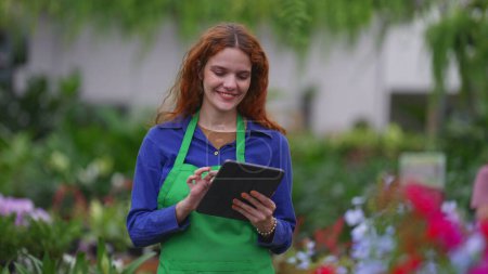 Téléchargez les photos : Joyeux fleuriste rousse jeune entrepreneur femme tenant la tablette et souriant à la caméra debout à l'intérieur du magasin de détail d'affaires - en image libre de droit