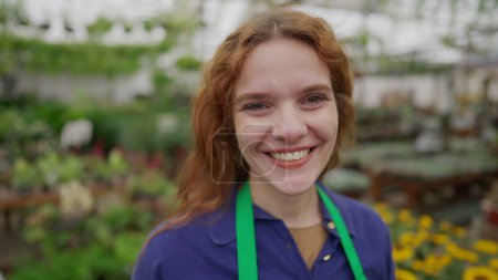 Foto de Retrato de una florista feliz De pie dentro de Flower Shop con delantal verde sonriendo a la cámara. Cara de cerca de una joven trabajadora alegre - Imagen libre de derechos