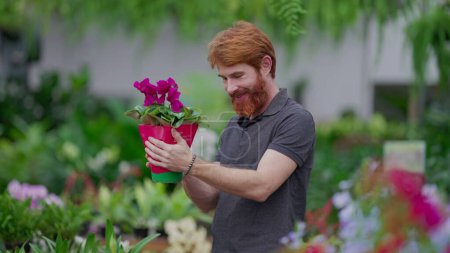 Foto de Un joven pelirrojo recogiendo flores de la estantería de pie dentro de la tienda de plantas de horticultura. Un hombre caucásico feliz comprando plantas - Imagen libre de derechos
