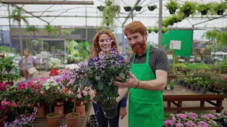 Photo for Man helping customer to select plant at Local Business Flower Shop. A redhead male person with green apron assisting a female customer - Royalty Free Image