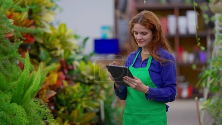 Photo for Joyful Female Manager Conducting Inventory Check in Plant Retail Store. Young Woman Utilizing Tablet while Navigating Business Shop Aisle - Royalty Free Image