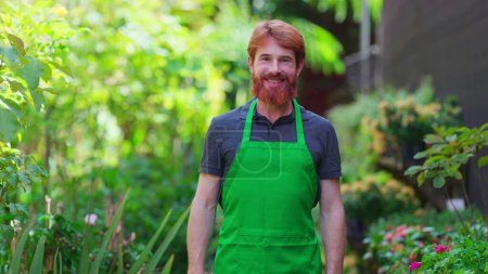 Photo for One happy young male gardener wearing Green Apron with arms crossed smiling at camera in natural garden environment - Royalty Free Image