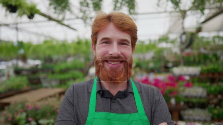 Photo for Young worker with arms crossed standing inside Flower Shop. A happy red hair male employee with green apron smiling at camera - Royalty Free Image