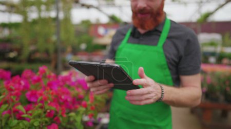 Photo for A male Staff using tablet to browse inventory inside Flower Shop. Young man wearing green apron holding modern technology - Royalty Free Image