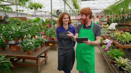 Photo for Two Staff colleagues walking through flower shop holding tablet. Happy redhaired man and woman wearing green apron inside plant local business store - Royalty Free Image