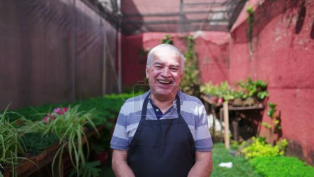 Foto de Retrato de un jardinero masculino mayor sonriendo a la cámara de pie en el patio trasero de Flower Shop. alegre persona mayor usando delantal en tienda de plantas locales - Imagen libre de derechos