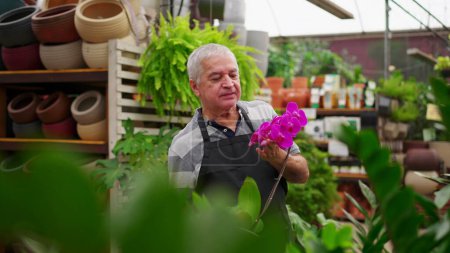 Foto de Retrato de un hombre mayor Florista empleado sosteniendo la flor dentro de la tienda de plantas hortícolas mientras gira la cabeza a la cámara sonriendo - Imagen libre de derechos