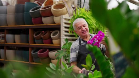 Foto de Retrato de un hombre mayor Florista empleado sosteniendo la flor dentro de la tienda de plantas hortícolas mientras gira la cabeza a la cámara sonriendo - Imagen libre de derechos