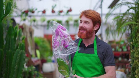 Téléchargez les photos : Heureux jeune homme employé de magasin de plantes portant des fleurs. Un homme rousse caucasien portant tablier vert marche à travers l'allée du magasin d'affaires - en image libre de droit